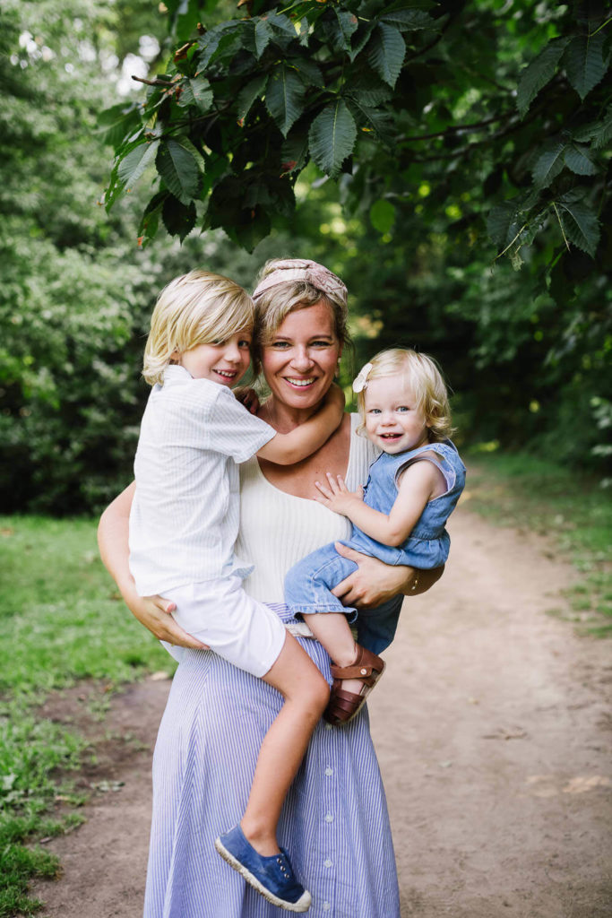 A woman in a white top and blue skirt stands on a forest path, happily holding two children, one in each arm. The older child wears a white shirt and shorts, while the younger wears a blue outfit. Green trees surround them.
