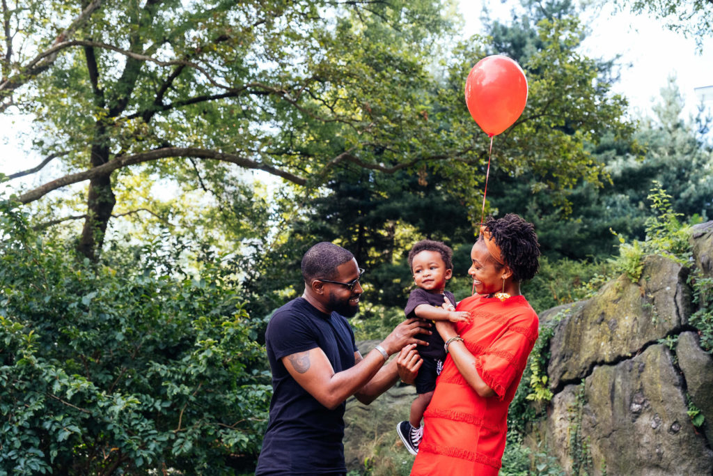 A smiling family stands outdoors in front of greenery and rocks. The father, wearing sunglasses and a black shirt, and the mother, in a red dress, hold their young child, who is grasping a red balloon. Trees and rocks form the background.