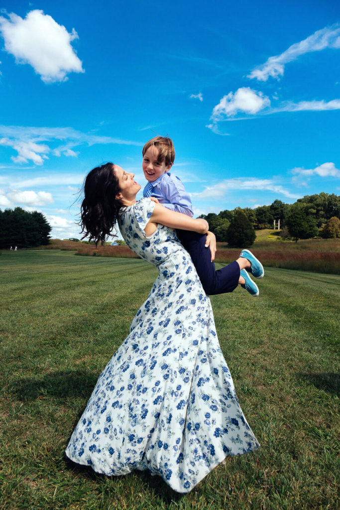 A woman in a flowing blue floral dress joyfully spins a smiling child in a grassy field under a bright blue sky with scattered clouds. Trees line the horizon in the background.
