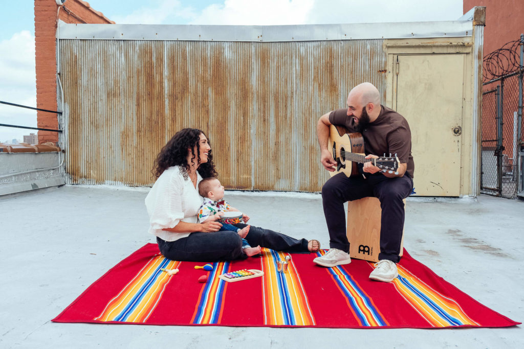 A family sits on a colorful striped blanket on a rooftop. A man plays guitar while a woman and a child sit nearby. The background features a corrugated metal wall and a door. The atmosphere is relaxed and joyful.
