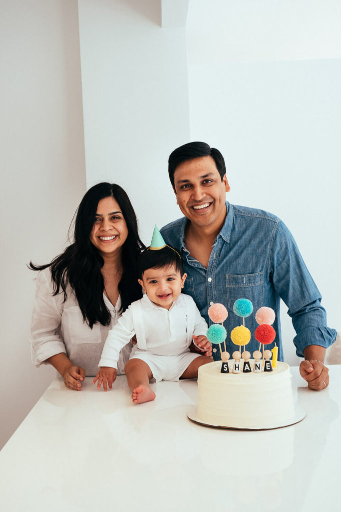 A smiling family stands around a birthday cake on a table. A baby wearing a paper party hat sits between two adults. The cake is decorated with colorful pom-pom toppers and letter candles spelling Shane.