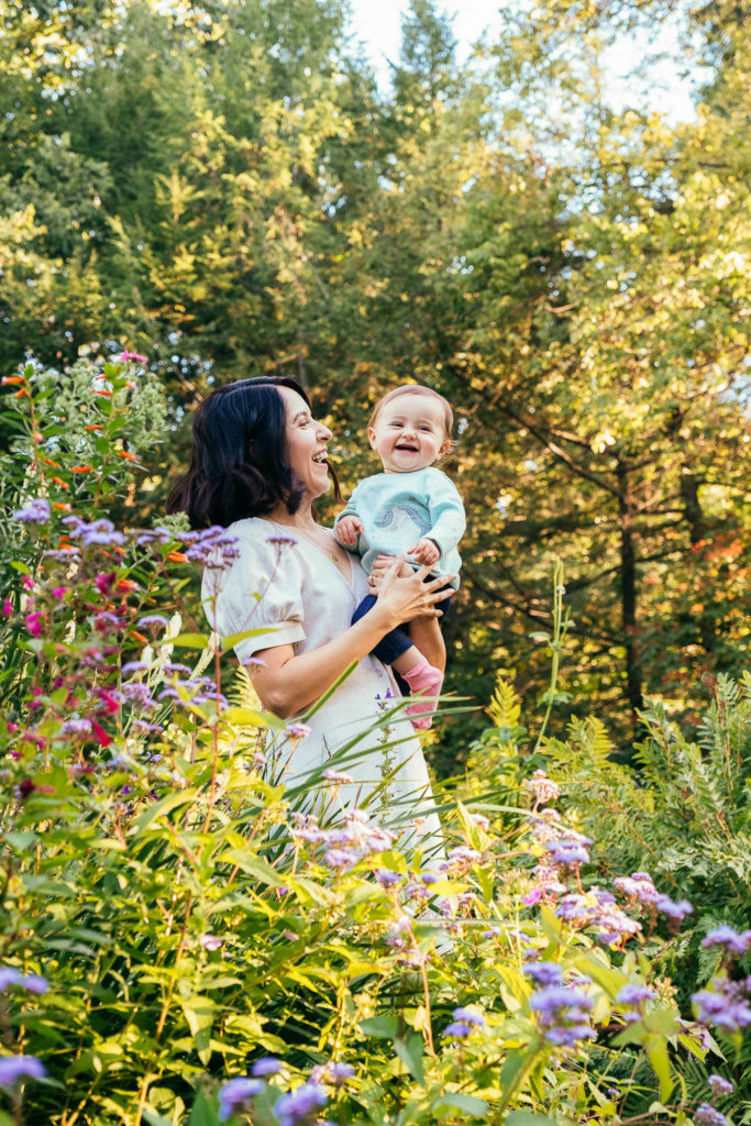A woman in a white dress holds a smiling baby in a vibrant, flower-filled garden. Sunlight filters through the trees, casting a warm glow over the scene. The woman and baby are surrounded by colorful wildflowers.
