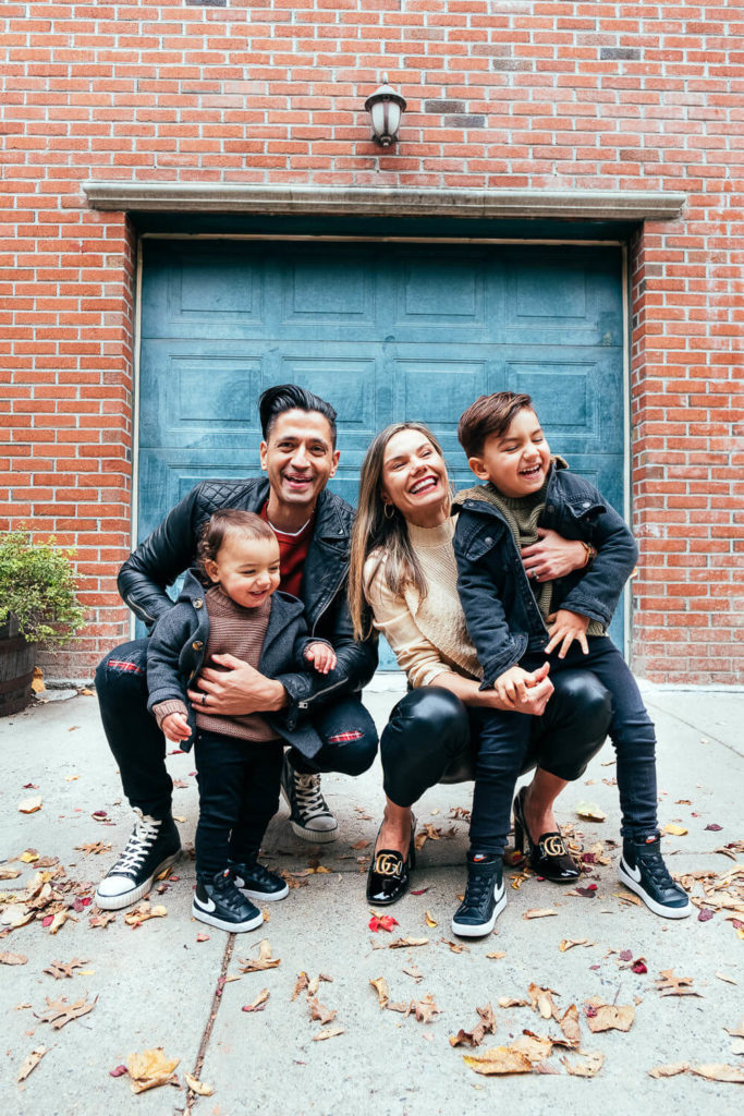 A family of four poses in front of a blue garage door. The parents, both wearing jackets, crouch down to embrace their two young children. The ground is scattered with autumn leaves, and everyone is smiling happily.