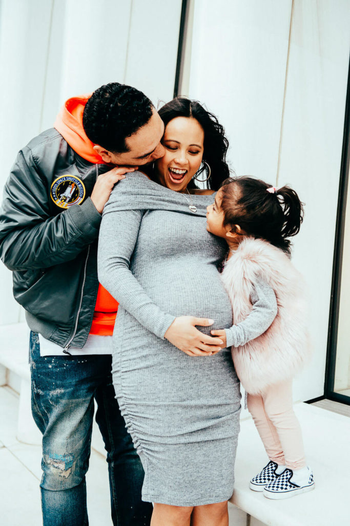 A joyful family scene: A man embraces a pregnant woman from behind, while a young girl leans in to kiss the womans belly. The woman wears a gray dress, the man a jacket with an orange hoodie, and the girl a pink vest. They are smiling brightly.