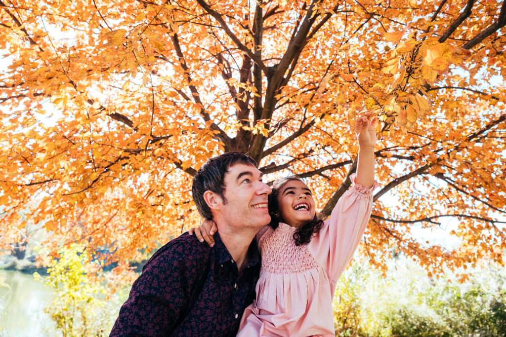 A man and a young girl are joyfully under a tree with vibrant orange leaves. The man is carrying the girl, who is reaching up towards the leaves, both smiling brightly. The background shows a sunny day in a park setting.