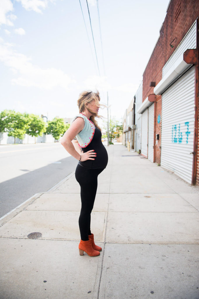 A pregnant woman stands confidently on a deserted sidewalk. Shes wearing a black outfit with a light blue vest and orange boots. Her hair is blowing in the breeze as she looks off into the distance. The street is lined with closed shops and trees.