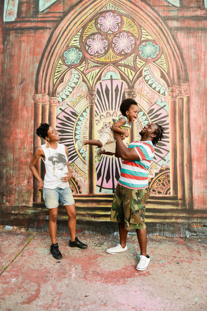 A joyful family scene in front of a colorful mural depicting a large, decorative arched window. One person playfully lifts a child in the air, while another watches and smiles. The background features vibrant patterns and details.