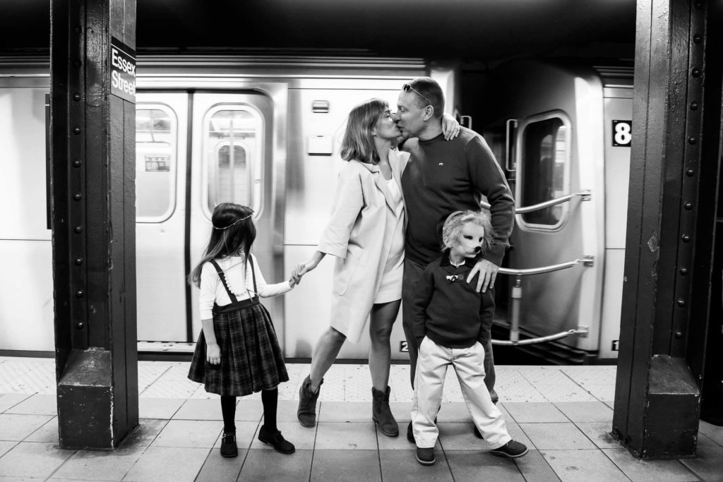 A black-and-white photo shows a family at a subway station. A couple kisses while their daughter stands holding the mothers hand, and the son stands beside the father. A train is passing in the background.