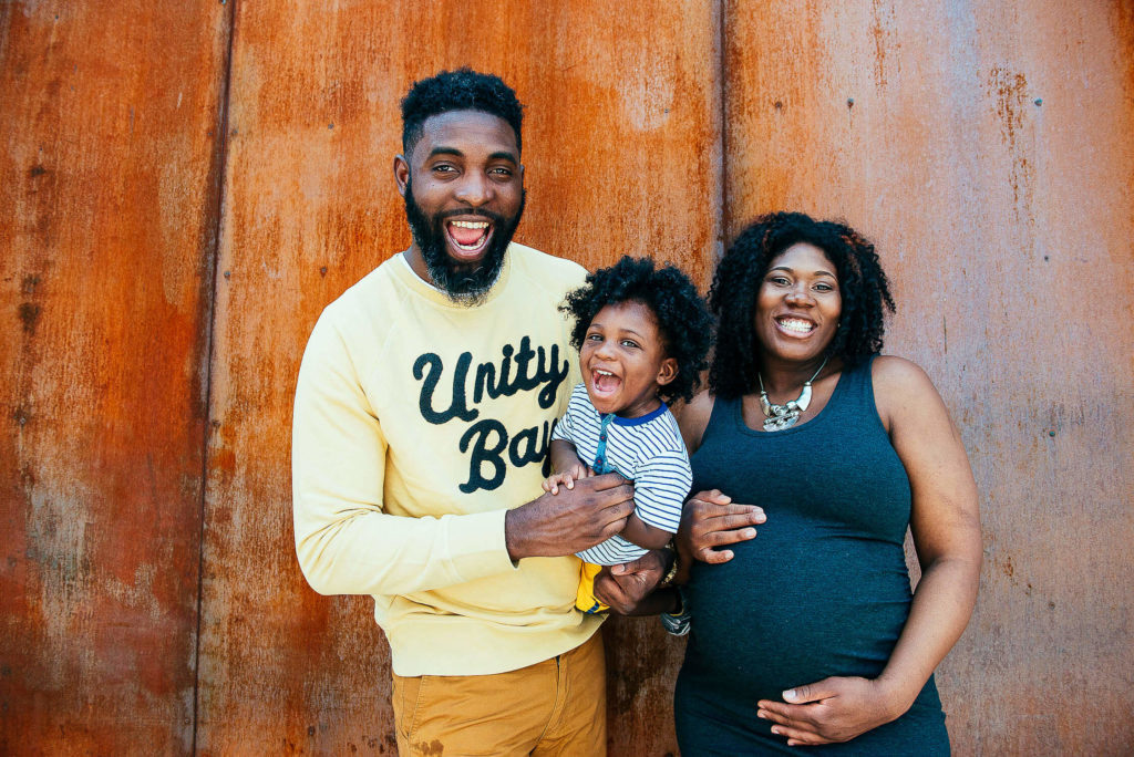 A joyful family stands in front of a rust-colored wall. A bearded man in a yellow sweater holds a laughing young child. A smiling pregnant woman in a black dress stands beside them, resting her hand on her belly.