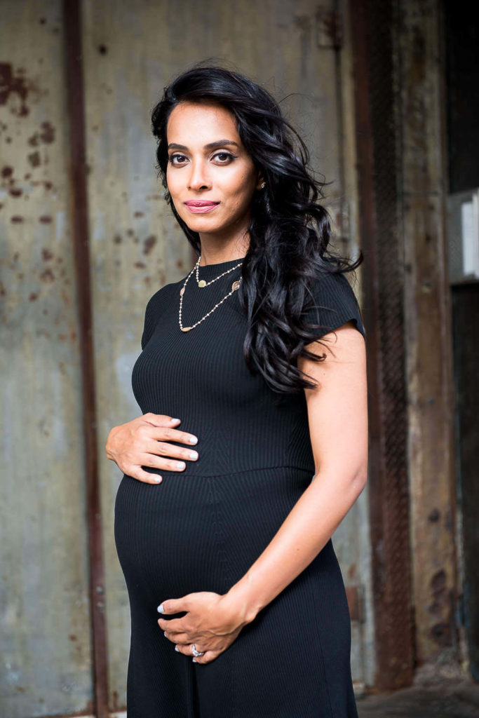 A woman in a black dress cradles her baby bump with both hands. She has long, wavy hair and stands against a rustic metal backdrop, wearing a gentle smile.