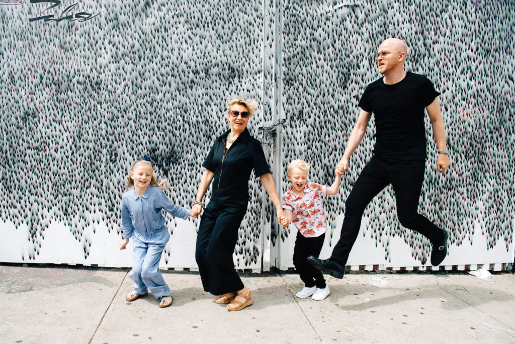 A family of four, two adults and two children, joyfully holding hands and jumping in front of a patterned gray and white wall. All are wearing casual clothes, and the children smile widely. The father is mid-jump, creating a playful scene.