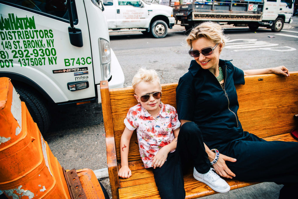 A woman and a child wearing sunglasses sit together on a wooden bench outdoors. The child is dressed in a floral shirt and white sneakers, while the woman wears a black outfit. Trucks are parked on the street in the background.