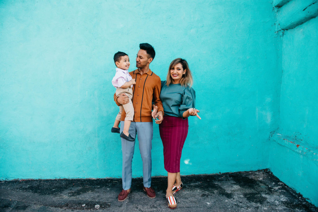 A family of three stands against a bright turquoise wall. The father holds a young child, and the mother stands beside them, smiling. They are all casually dressed, with the father in a brown shirt and gray pants, and the mother in a teal top and red skirt.