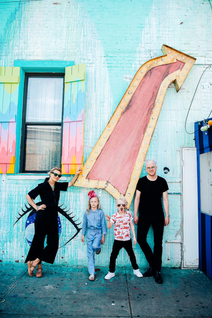 A family of four poses in front of a colorful, artistic mural featuring a large arrow and eye design. The mother wears a black outfit, while the father and two children wear casual clothes. The setting is vibrant and urban.