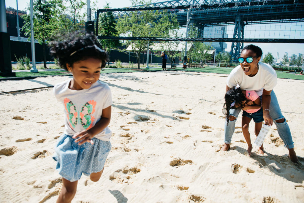 A woman and two young children play joyfully on a sandy volleyball court. One child runs towards the camera, while the woman playfully holds the other child. A bridge and trees are visible in the background.