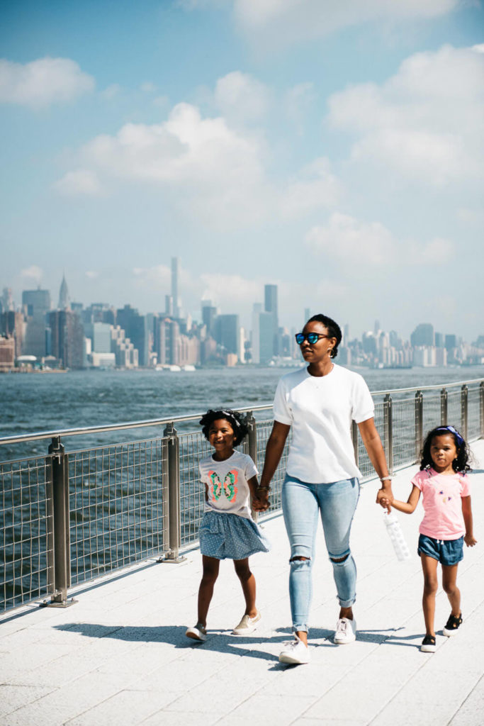 A woman walks with two children along a waterfront promenade on a sunny day. The background features a city skyline with skyscrapers and a blue sky with clouds. The woman wears sunglasses, a white t-shirt, and jeans. The children wear colorful outfits.