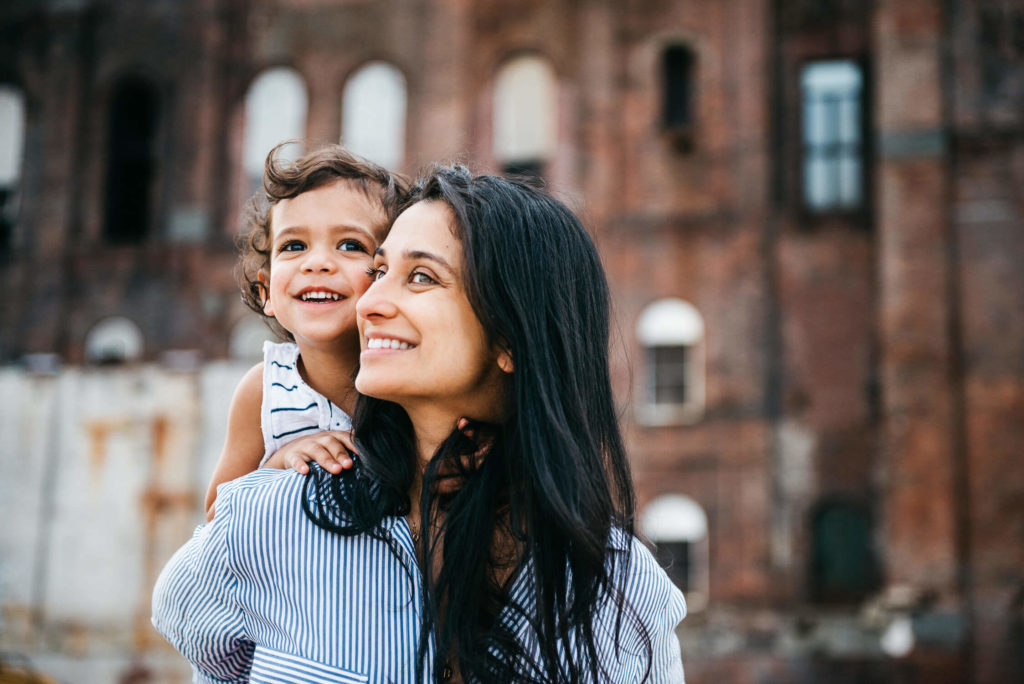 A woman carrying a smiling child on her shoulders stands in front of a rustic brick building. The woman looks up, and both appear happy and content. The background reveals old architecture with several windows.