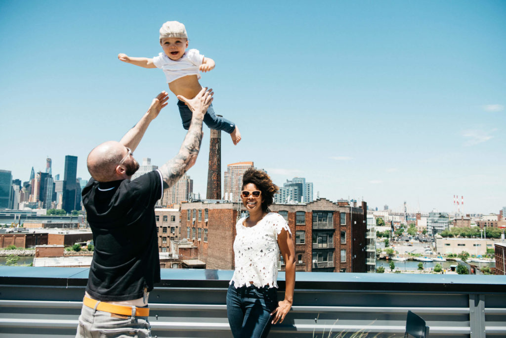 A man playfully tosses a toddler in the air on a rooftop with a city skyline in the background. A woman stands nearby, smiling. The sky is clear and blue.