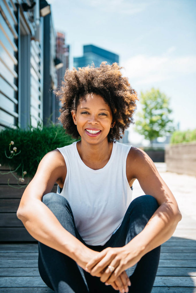 A person with curly hair in a white sleeveless top and dark pants sits outdoors, smiling. They are leaning forward with their hands resting on their knees. The background features a building, greenery, and a clear sky.