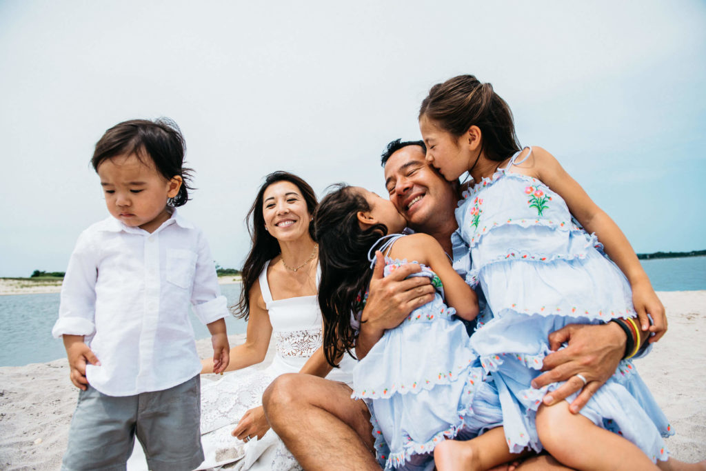 A family sits on a sandy beach. A man is smiling as two young girls in matching blue dresses kiss his cheeks. A woman sits smiling beside them, and a young boy in a white shirt stands to the side. The background features a calm body of water.