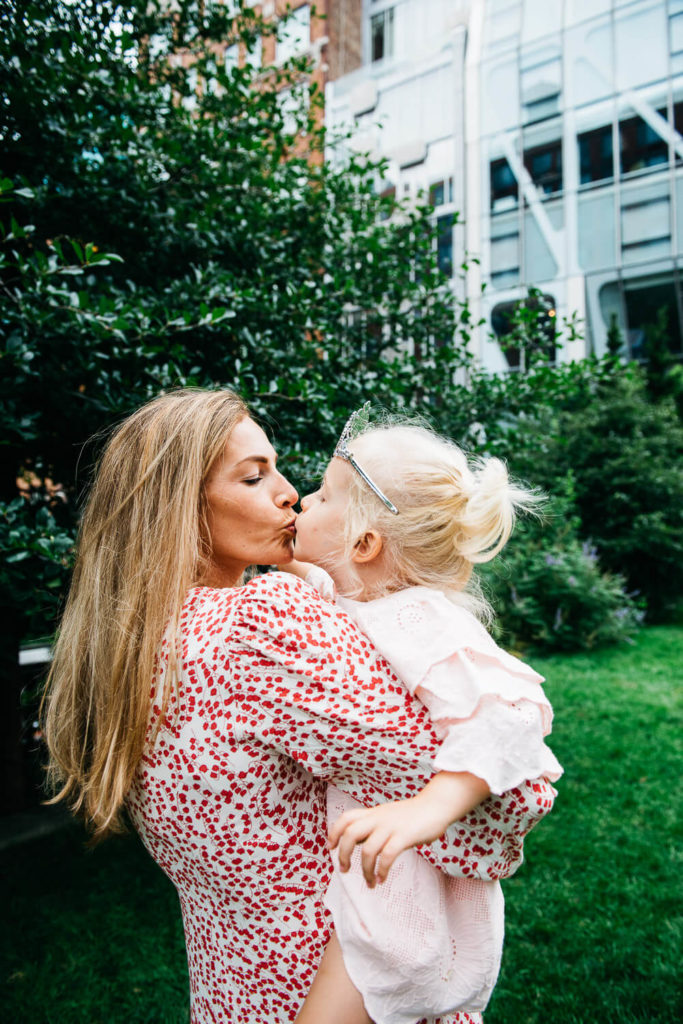 A woman kisses a young girl outdoors. The woman wears a red and white patterned dress, and the girl, with a silver tiara, wears a light pink dress. They are surrounded by greenery with a building in the background.
