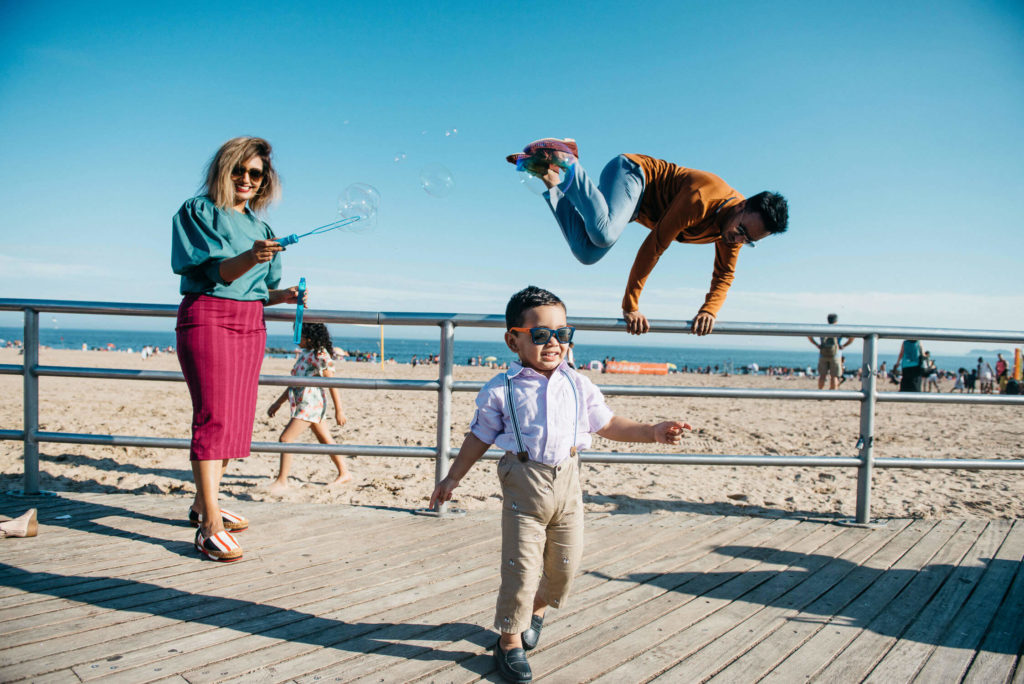 A stylish woman in sunglasses blows bubbles on a boardwalk. A young boy in sunglasses walks confidently nearby. A man playfully leaps over a railing in the background with a beach scene behind them.