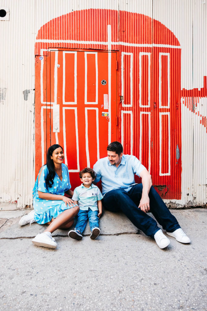 A smiling family of three sits on the ground in front of a red and white painted wall. The parents and their young child are casually dressed in blue outfits. The wall design resembles a door with abstract patterns.