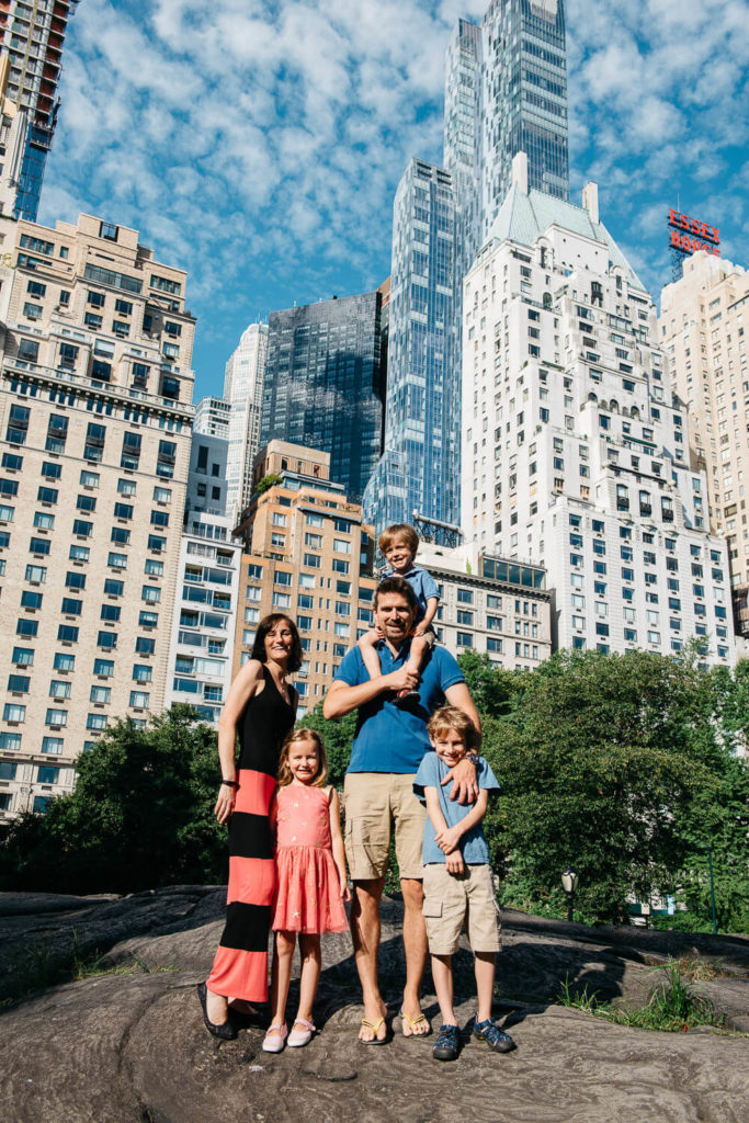 A family of five stands on a rock in a city park. The father carries a child on his shoulders, while the mother and two other children stand by his side. Tall buildings and a cloudy blue sky are visible in the background.