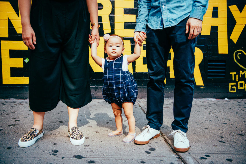 A child in a blue checkered outfit holds hands with two adults on a sidewalk. The adults wear casual shoes and clothing. The background features a colorful wall with yellow text.