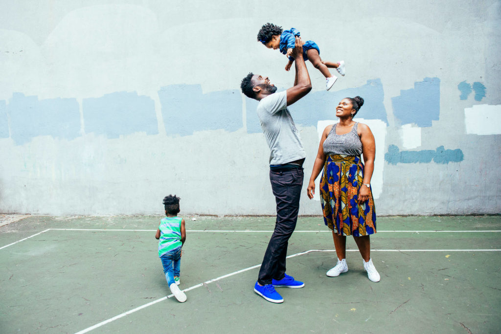 A family enjoys time together on a basketball court. The father, wearing a gray shirt and blue sneakers, lifts a young child into the air. The mother, in a patterned skirt, smiles while another child in green stands nearby.