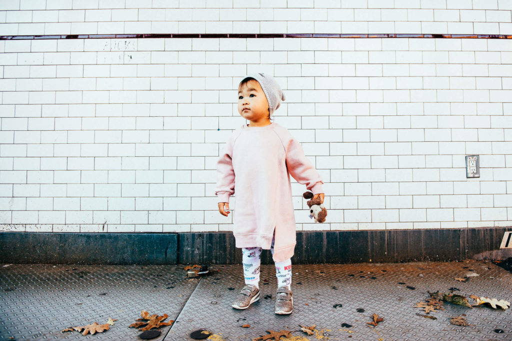 toddler in a pink outfit standing against a white brick wall with leaves on the ground