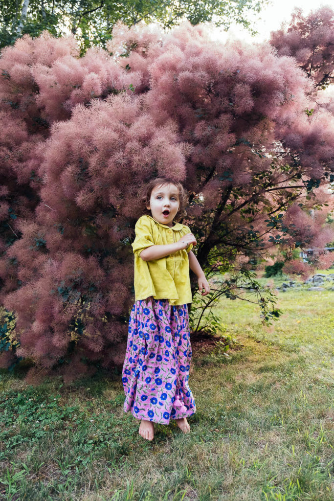 surprised girl in a bright yellow top and colorful skirt standing in front of a tree with fluffy pink foliage