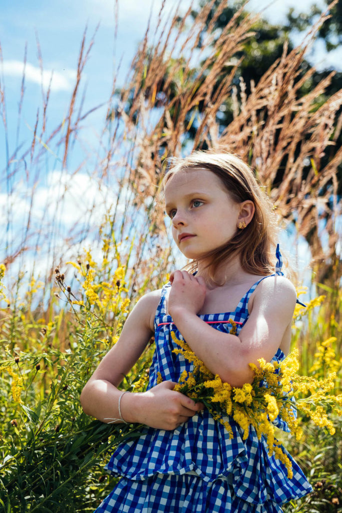 Young girl in a blue checkered dress holding yellow wildflowers in a grassy field.