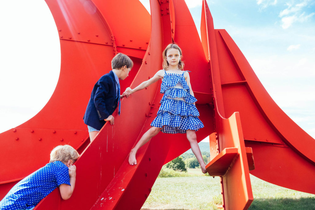 girl climbing a large red sculpture with friends nearby, exploring outdoor art in a blue summer dress