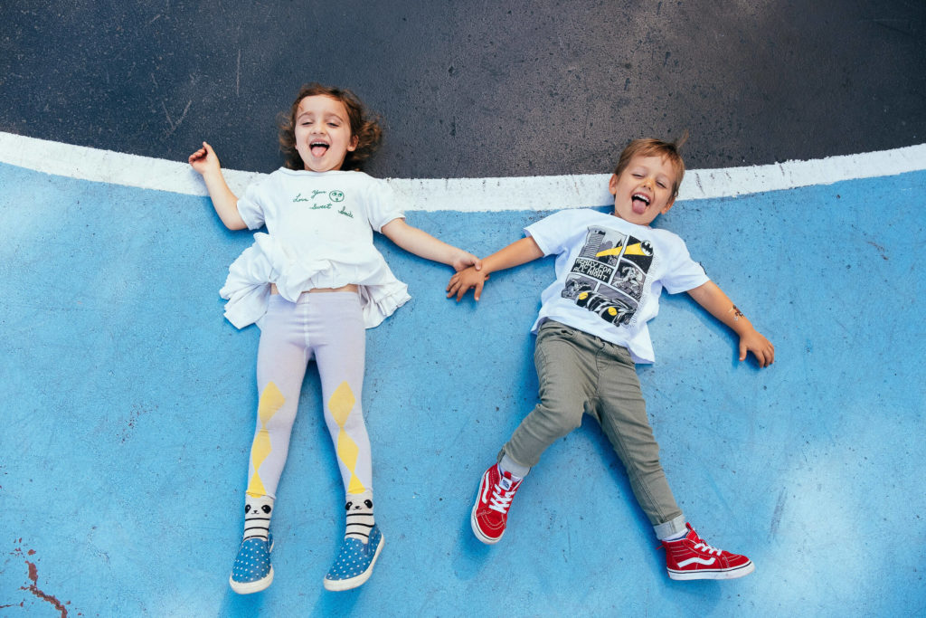 two children laughing while lying on a blue playground surface, holding hands