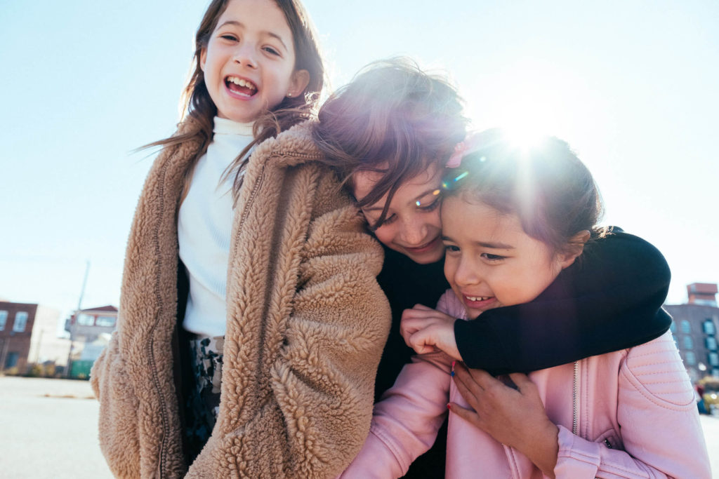 Girl standing outdoors with a wind-swept coat, looking toward the distance against a metal building and blue sky.
