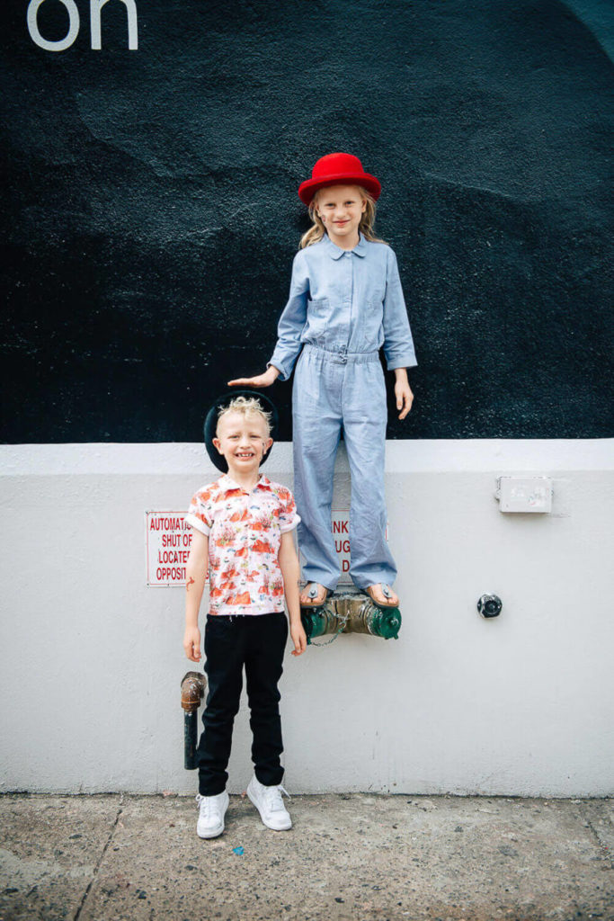 two kids with playful expressions; one in a red hat standing on a ledge, other in casual clothes below