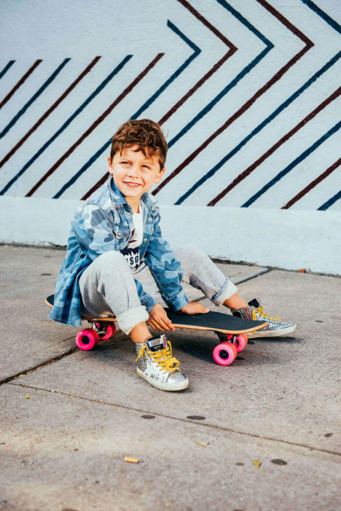 young boy smiling while sitting on a skateboard in a denim jacket and yellow sneakers, urban background