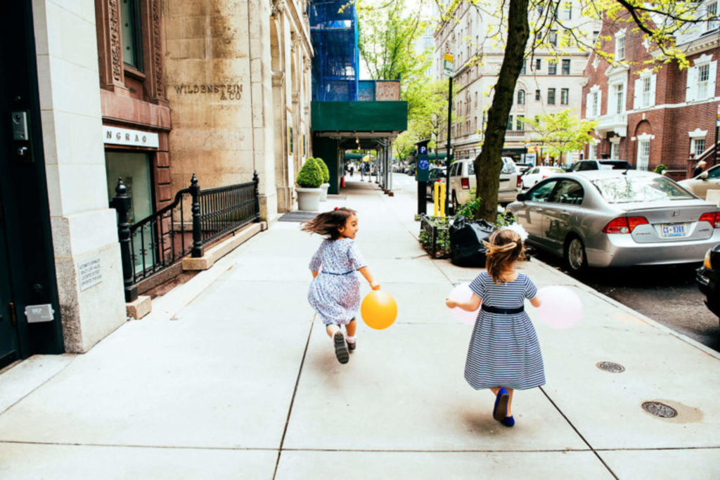 two young girls with balloons, joyfully running down a city sidewalk