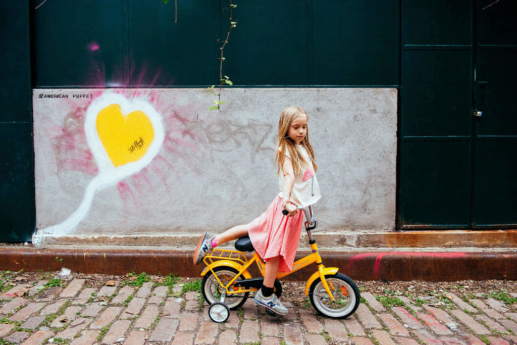 Stylish young girl posing with a yellow bike and heart graffiti backdrop in Brooklyn.