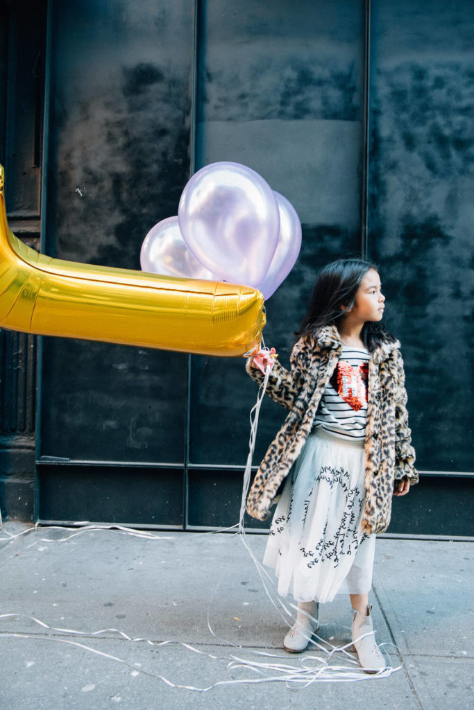 girl in a leopard print coat holding yellow and purple balloons, standing on a city street