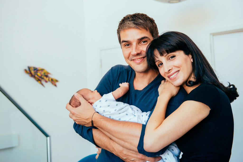 A couple smiling and holding a newborn baby wrapped in a patterned blanket. They stand in a brightly lit room with modern decor visible in the background.