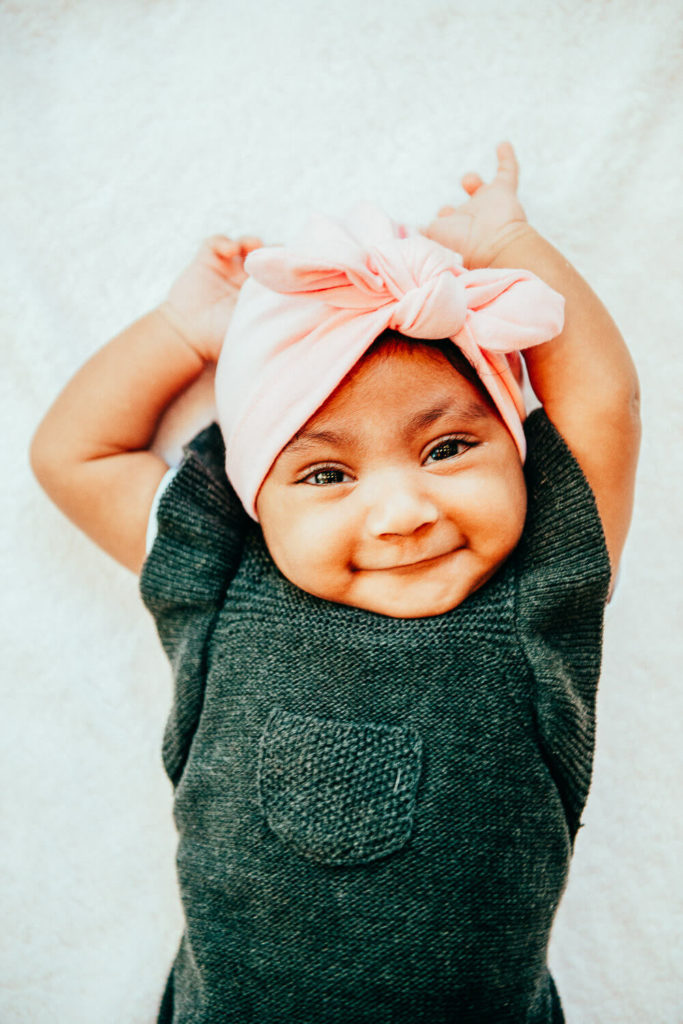 A smiling baby wearing a gray outfit and a pink headband with a bow lies on a white blanket, with arms stretched above their head.