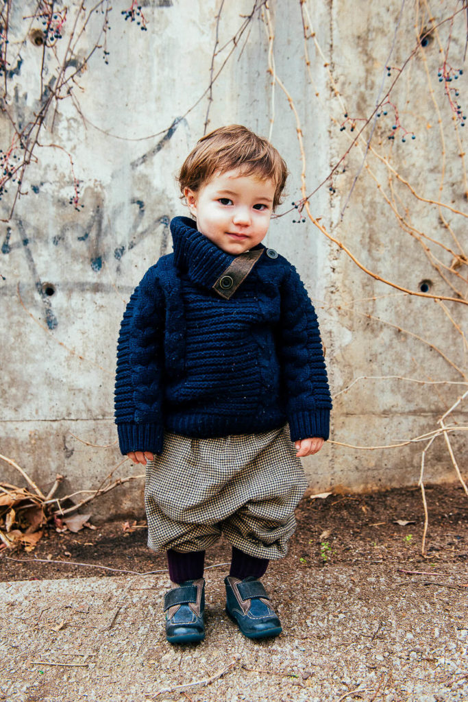 toddler in a cozy sweater and patterned pants, standing against a textured wall outdoors
