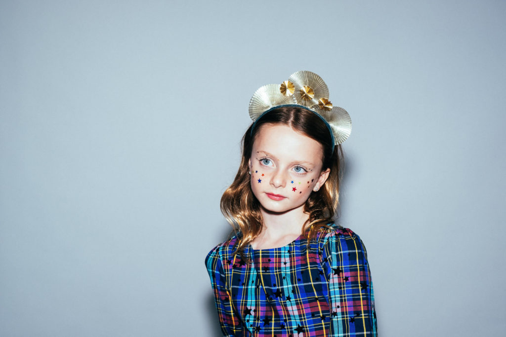 A child with wavy hair in a colorful plaid dress and starry makeup poses for a Brooklyn children’s portrait photographer. A paper-like crown adorned with gold stars sits atop their head, set against a plain gray background.
