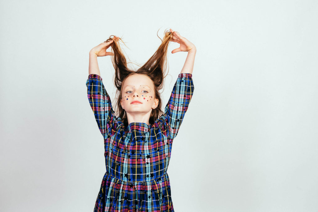 A young girl in a plaid dress playfully holds her long hair up with both hands against a plain backdrop. Captured by a Brooklyn children’s portrait photographer, she has colorful dots painted on her face and smiles confidently at the camera, showcasing the charm of children's portrait photography.