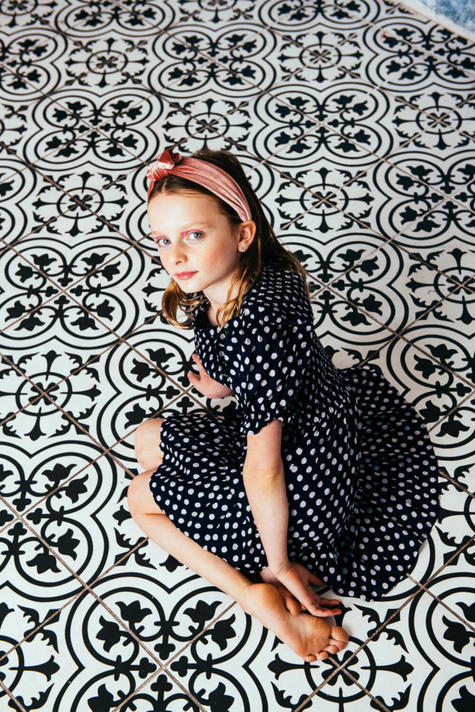 A young girl with long hair and a pink headband sits gracefully on a decorative black-and-white patterned floor. Wearing a black dress with white polka dots, she looks up at the camera, capturing the essence of children's portrait photography by a Brooklyn children’s portrait photographer.