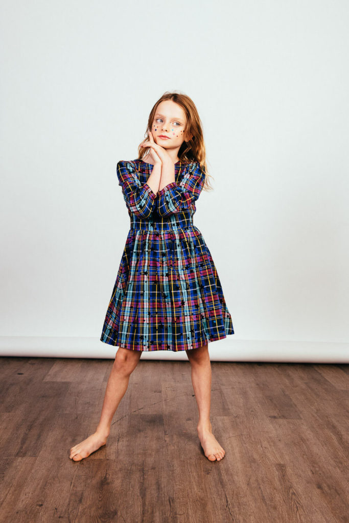 A young girl strikes a playful pose, standing barefoot on a wooden floor. She wears glasses and a colorful plaid dress. The backdrop is plain white, creating a simple and cheerful setting ideal for Brooklyn children's portrait photography.