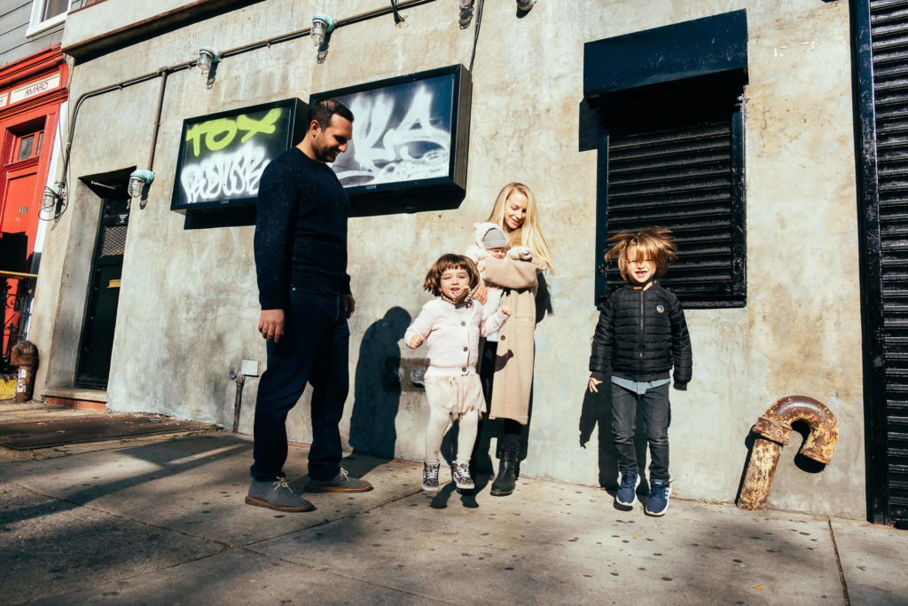 A family of four stands on a sunny Brooklyn street in front of a graffiti-covered wall. The mother cradles a baby, while the two older kids stand beside their father, all smiles in their warm clothing—a perfect moment captured by a talented family photographer.