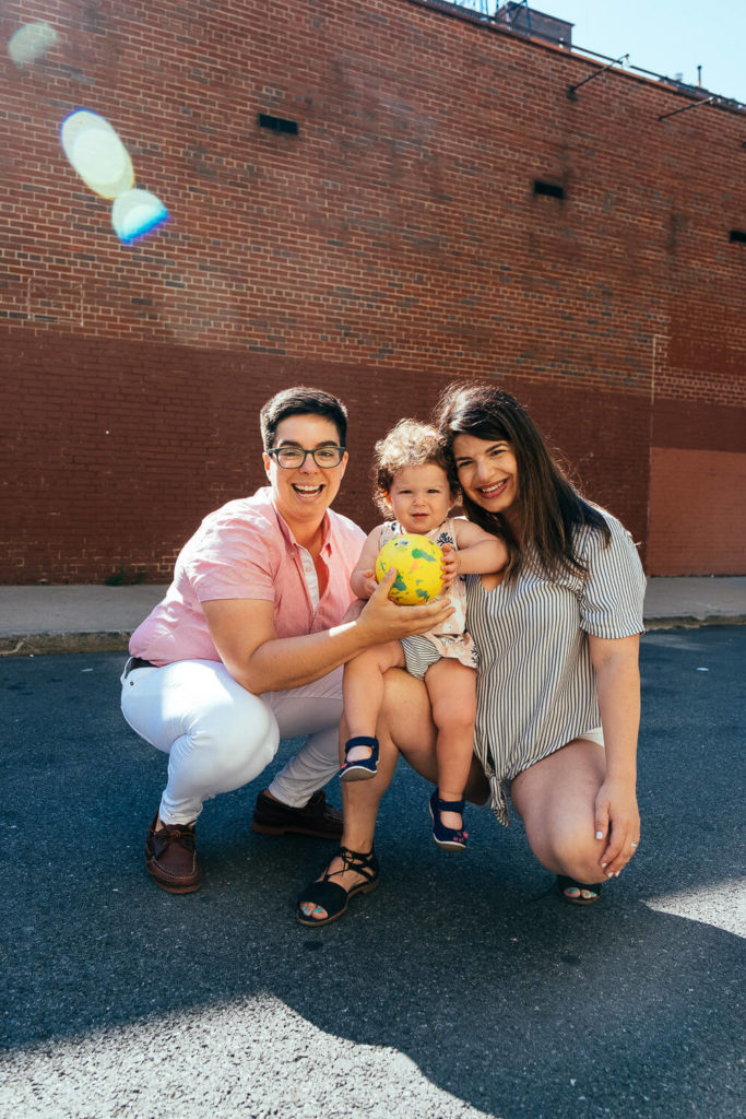 Two adults crouch beside a child holding a colorful ball on a sunny street, embodying the vibrant essence of family photography NYC. One adult wears a pink shirt and white pants, the other a striped outfit. They are all smiling, with a red brick wall in the background.
